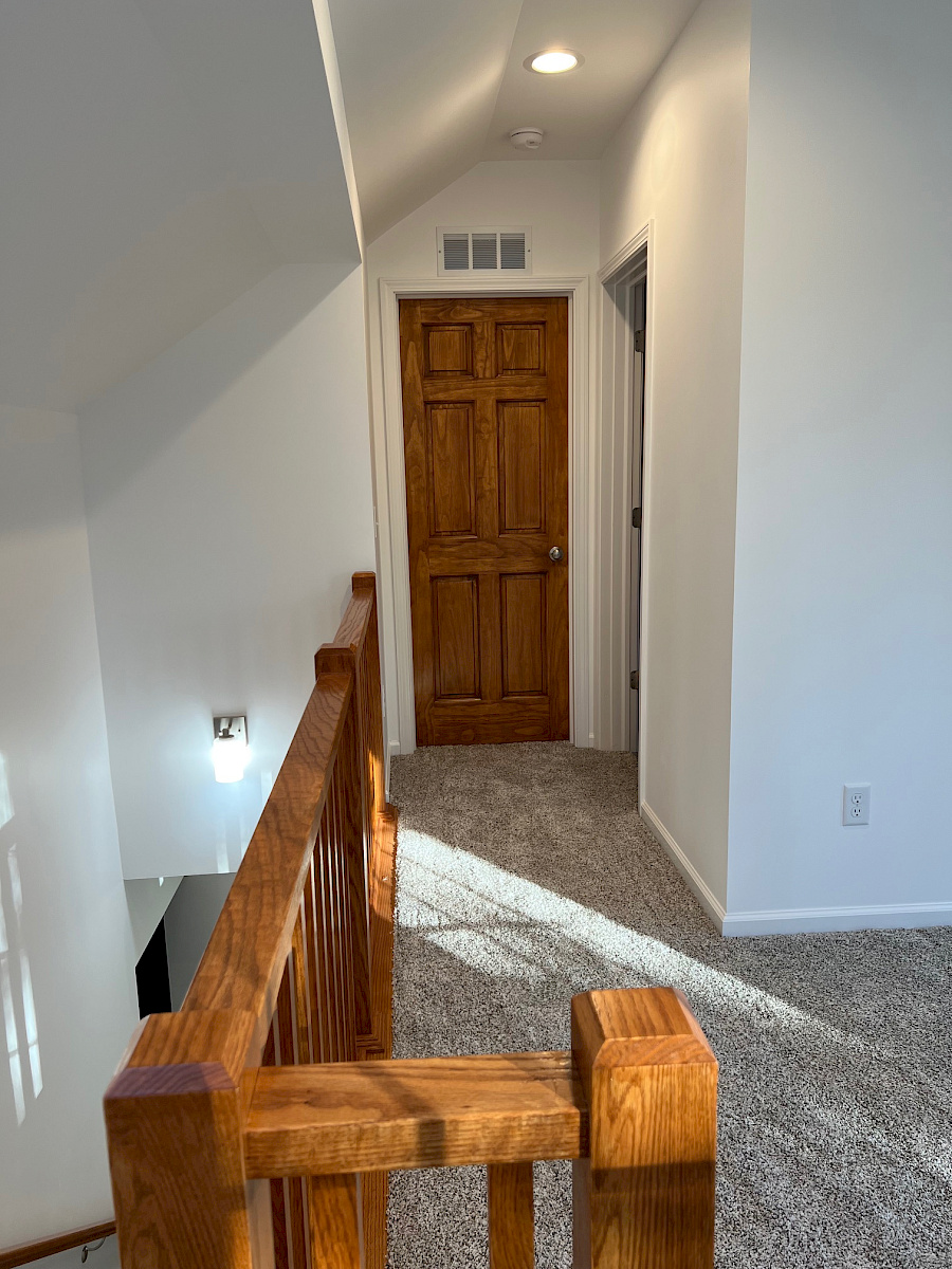 Second-floor landing with a stained six-panel door, vaulted ceiling, recessed lighting, and an oak railing.