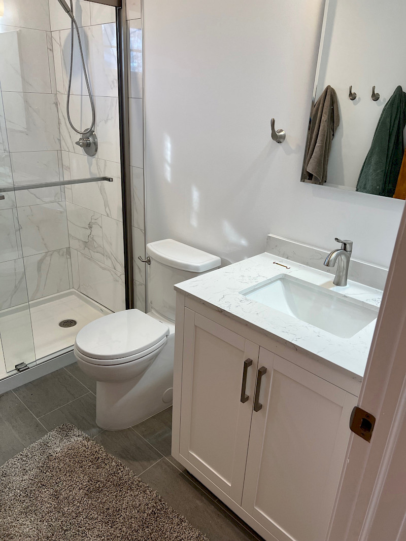 Modern bathroom remodel with a marble-look tiled walk-in shower, frameless glass door, white vanity, and quartz countertop.