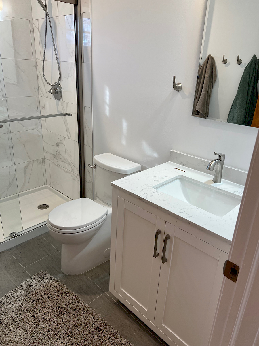 Modern bathroom remodel with a marble-look tiled walk-in shower, frameless glass door, white vanity, and quartz countertop.