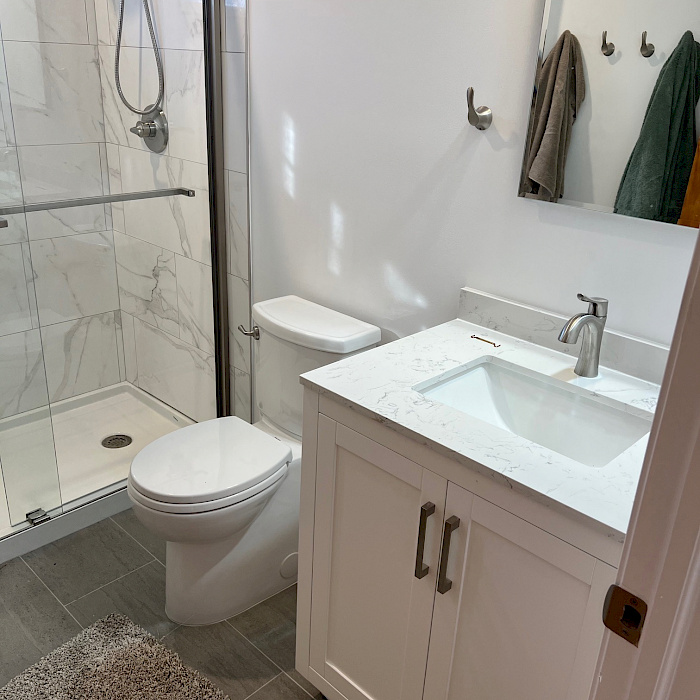 Modern bathroom remodel with a marble-look tiled walk-in shower, frameless glass door, white vanity, and quartz countertop.