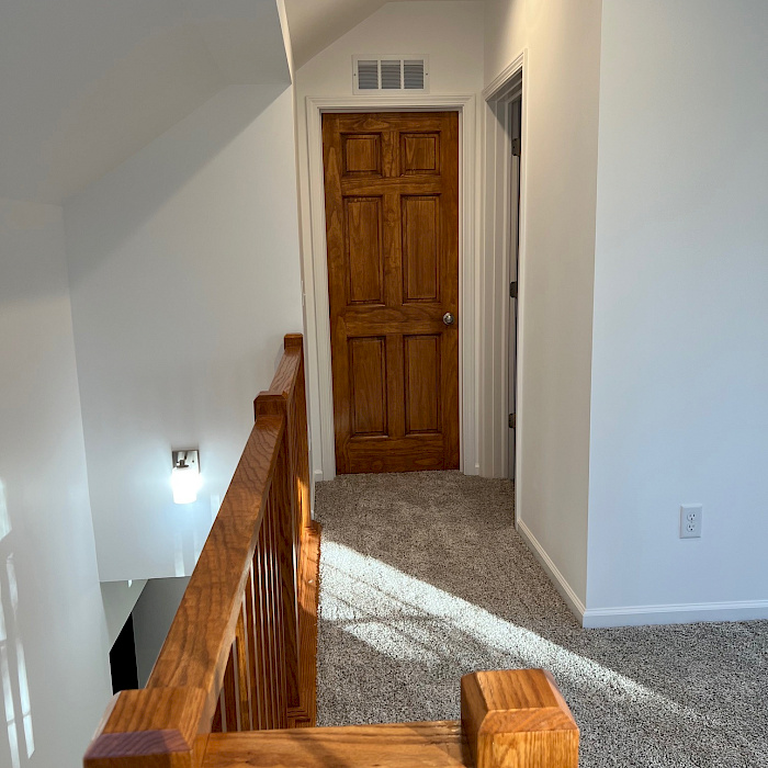 Second-floor landing with a stained six-panel door, vaulted ceiling, recessed lighting, and an oak railing.